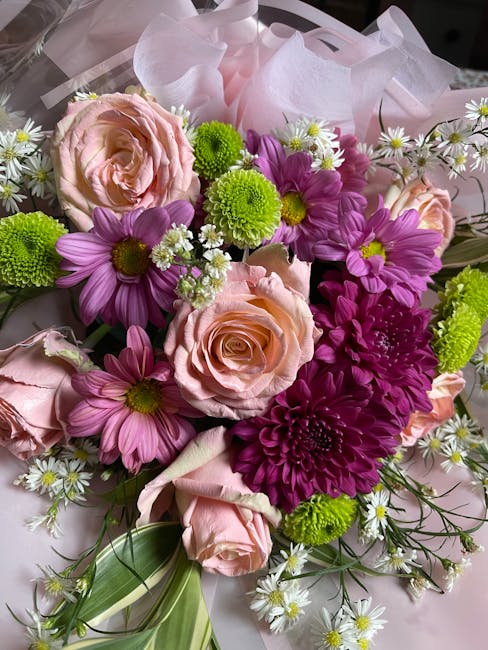 A close-up of a floral bouquet featuring soft pink roses with layered petals, vibrant purple daisies with yellow centers, bright green button chrysanthemums, and small white daisies with yellow center