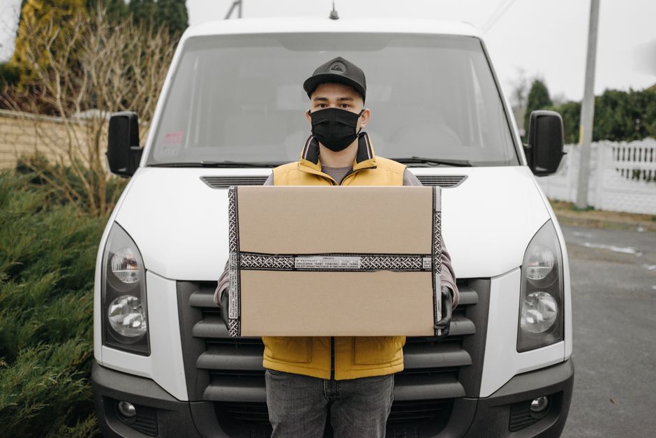 A delivery person wearing a black face mask, yellow vest, and dark cap standing outdoors in front of a white delivery van, holding a large rectangular floral gift box wrapped in beige paper with black