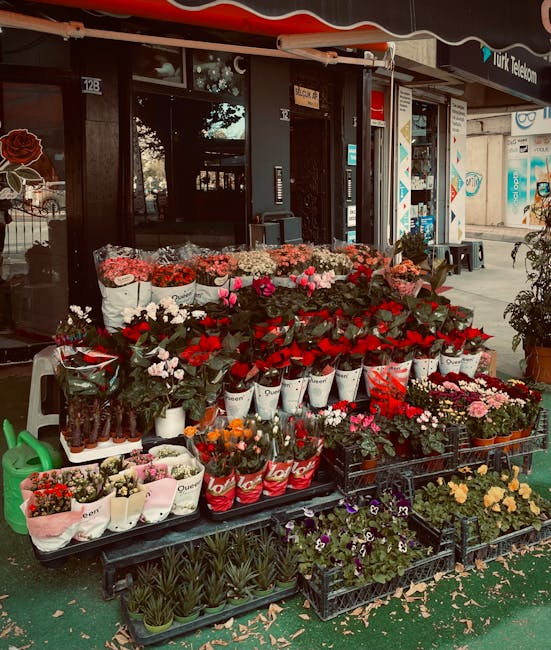 A vibrant floral display outside a flower shop, featuring a variety of fresh flowers arranged in buckets and crates. Prominent in the arrangement are red roses, pink tulips, white carnations, and mixe