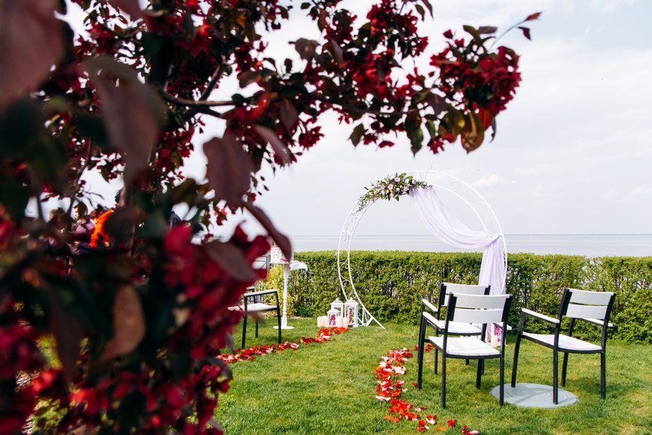 A scenic outdoor wedding setup with a circular floral arch decorated with white fabric and green foliage, positioned on lush green grass overlooking the sea. Four black and white chairs are arranged f