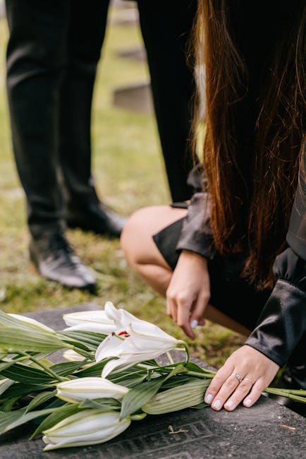 A person kneeling beside a memorial site, placing a floral tribute consisting of large white lilies, with long green leaves, on a black stone. The lilies have delicate, slightly curved petals and prom