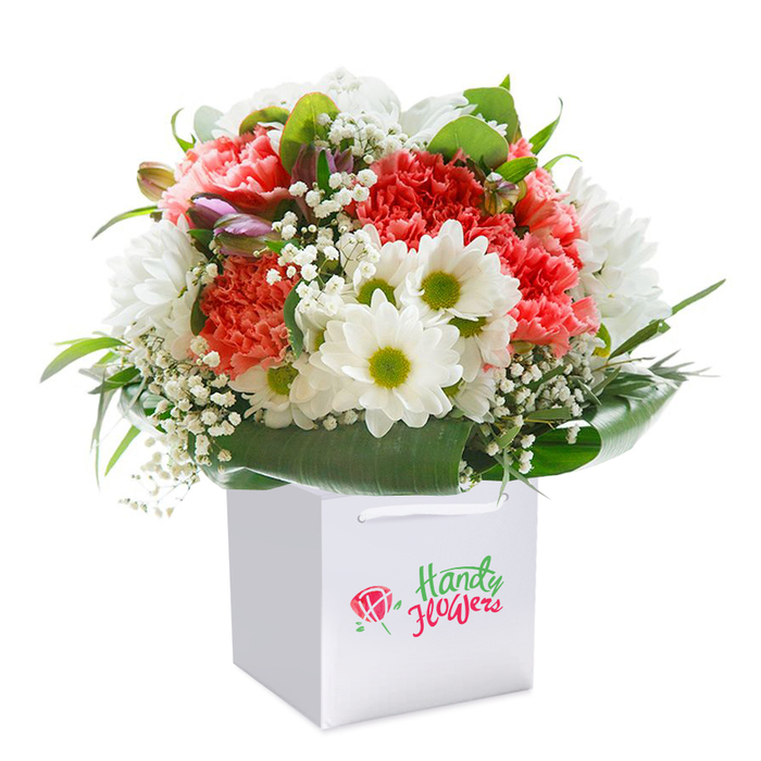 Perfectly poised for gifting and admiration, this luxuriously full floral arrangement is the work of skilled florists from the heart of Archway. The bouquet is meticulously designed, standing upright within a crisp, high-quality white square gift bag, whose soft ribbon handles glimmer gently under soft, natural lighting. The composition celebrates seasonal beauty: plush coral-pink and deep red carnations form a rich, textured heart, every petal brimming with youthful freshness. Framing these are bright white daisy-like blooms, their vibrant yellow-green centers echoing the cheerful parks and gardens scattered throughout the Archway area. Scattered in elegant proportion, delicate tufts of white baby's breath bring an ethereal softness, while a few purple-flushed buds tucked in on one side promise evolving colour and texture with each passing day. The deep, glossy green leaves, broad and polished, envelope the bouquet in a luxurious embrace, providing structure and a sense of natural abundance. The chic presentation in a sturdy gift bag not only ensures convenient delivery across Archway, from the historic Highgate Hill to the lively Market Square, but also enhances the delight of unwrapping a floral gift with a sense of anticipation. This bouquet, with its mix of soft hues and bold green foliage, communicates both contemporary elegance and the warmth of giving, making it an ideal choice for birthdays, anniversaries, or simply to add a touch of Archway cheer to any setting.