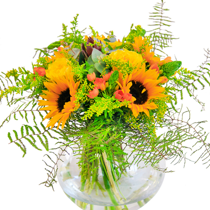This vibrant bouquet immediately captures the spirit of Archway's bustling main streets, bursting with the golden brightness of large sunflowers whose deep brown centers suggest contentment and grounded joy. Alongside, classic yellow garden roses lover's spiral outwards, each layered petal a testament to the area's blend of tradition and modernity. Sprays of glossy, scarlet Hypericum berries punctuate the arrangement, catching the eye like flashes of color along Junction Road. Wispy ferns and light, airy filler greens tumble gently over the edge, their movement evoking breezes through the Archway Bridge trees. Eucalyptus leaves in subtle blue-green shades peek amid the brighter hues, lending calm and sophistication. At the base, a round fishbowl-styled vase, filled with crystal-clear water, displays a web of vibrant stems-each slender line contributing to the bouquet's natural harmony. Up close, one can appreciate the tactile contrasts: velvety sunflower heads, tightly furled rose petals, and the crisp texture of fresh foliage. The bouquet exudes a light, uplifting fragrance, recalling the scent of cultivated gardens and summer air wafting from Elthorne Park. Designed as an uplifting centerpiece or heartfelt gift-whether for a celebration near Girdlestone Road or a simple moment of gratitude-this arrangement, curated with Archway sensibility and expertise, radiates happiness and connection.